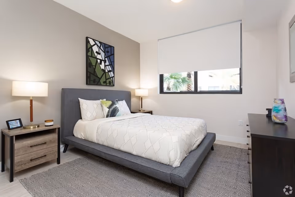 Modern bedroom with a gray upholstered bed, bedside tables and lamps, a dresser, and a window with a roller shade.