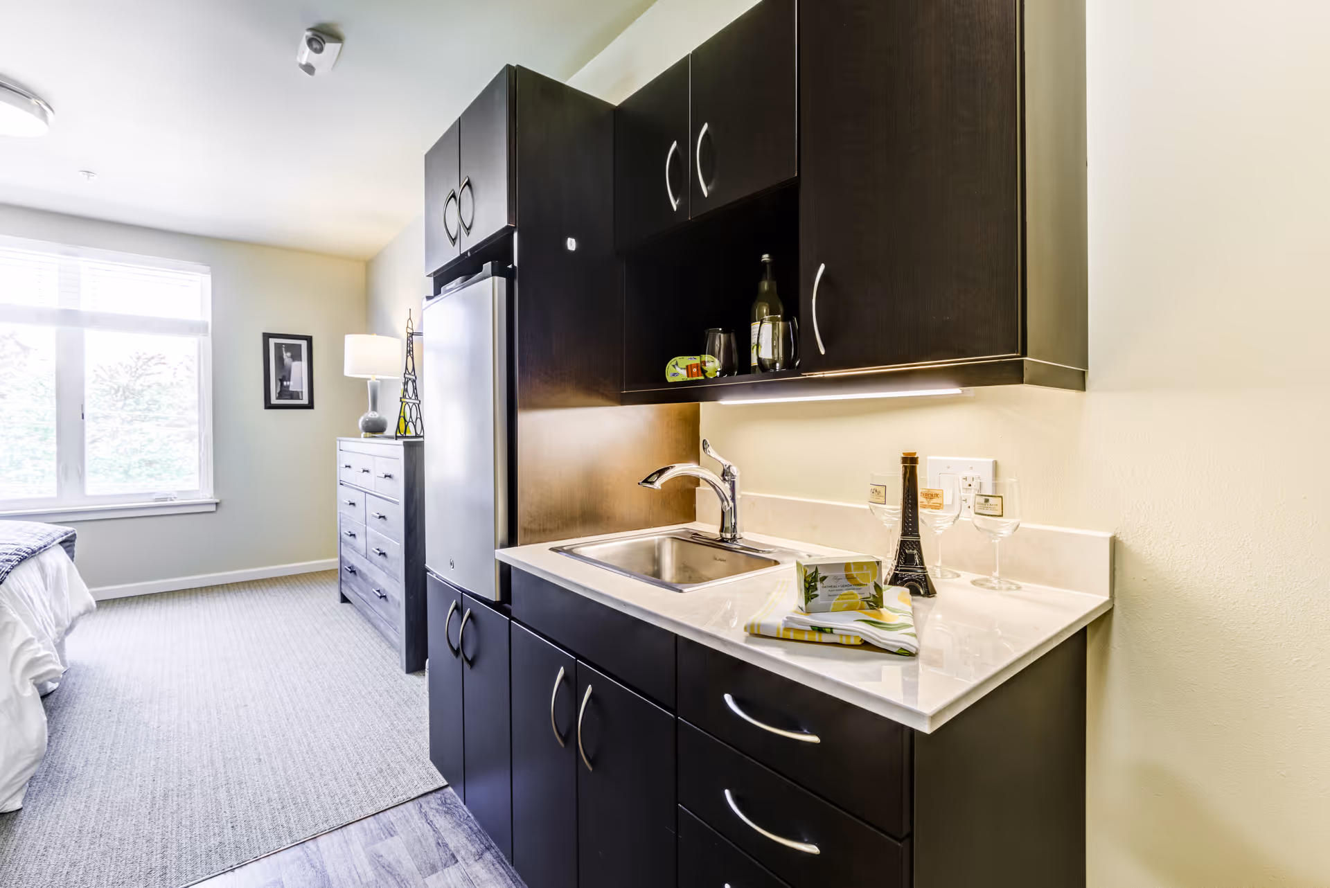 A small kitchenette area with dark wood cabinets, a sink, and a countertop with a bottle, two wine glasses, and a folded napkin. In the background, there is a window with blinds, a dresser with a lamp and decorative items, and part of a bed visible in a well-lit room.
