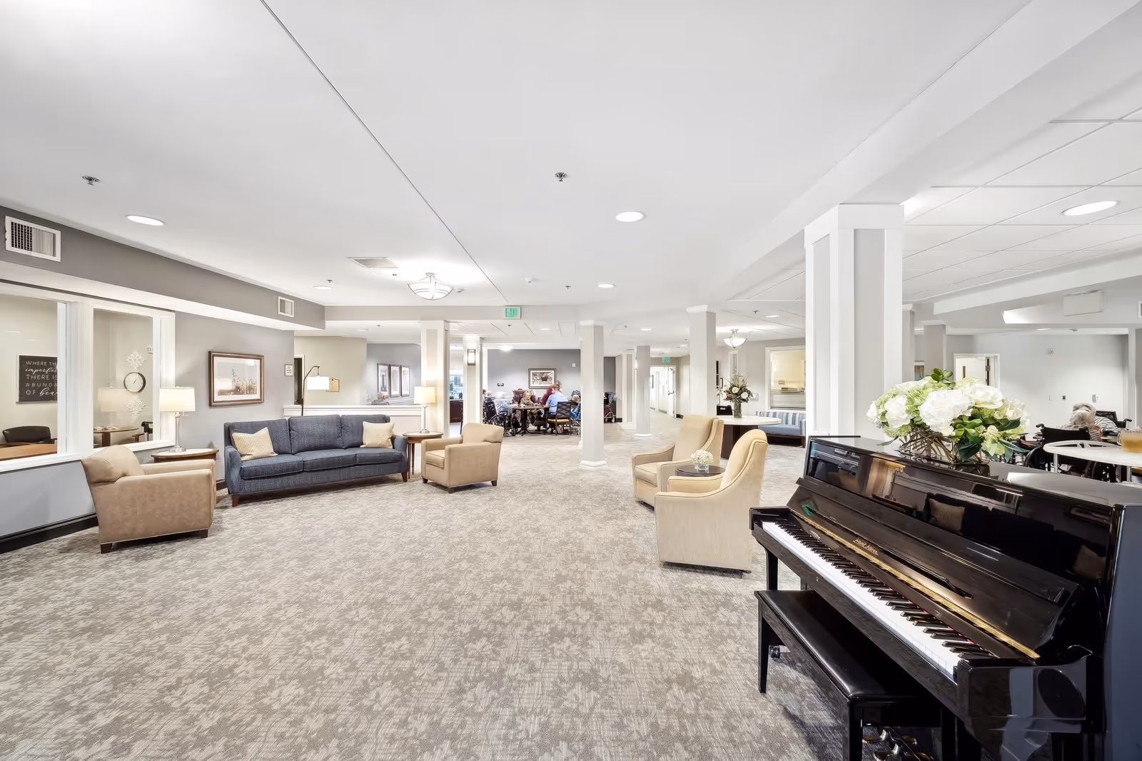 Spacious senior living facility common area with a piano on the right, beige armchairs, a blue sofa, and additional seating. The room is well-lit with ceiling lights and lamps, and there are people seated at tables in the background.