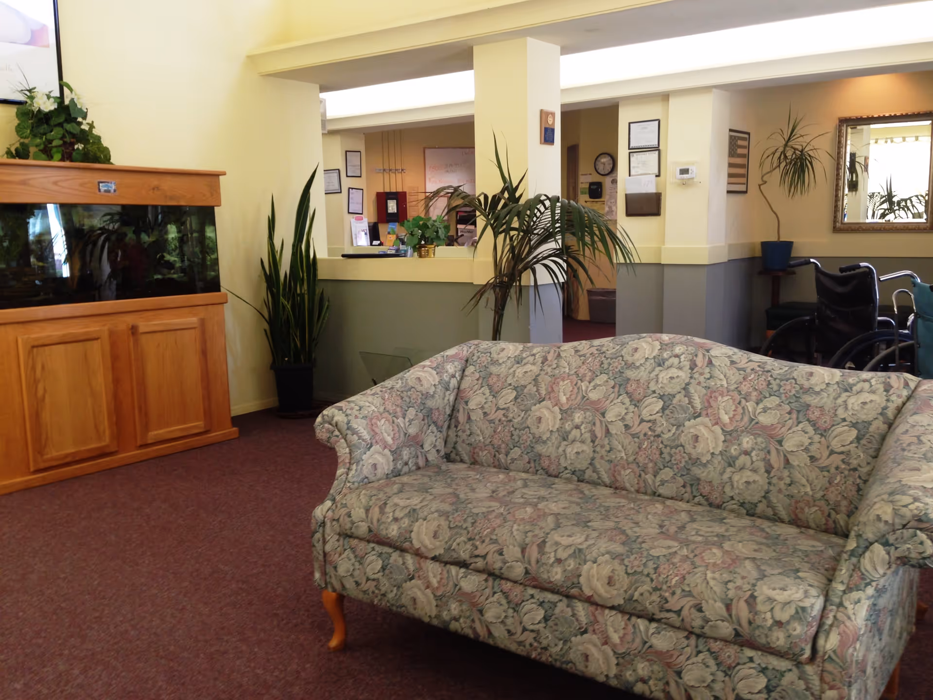 A cozy assisted living facility common area with a floral patterned sofa in the foreground, an aquarium with wooden cabinetry on the left, several potted plants, a reception desk in the background, and a wheelchair near a wall with a mirror and framed pictures.