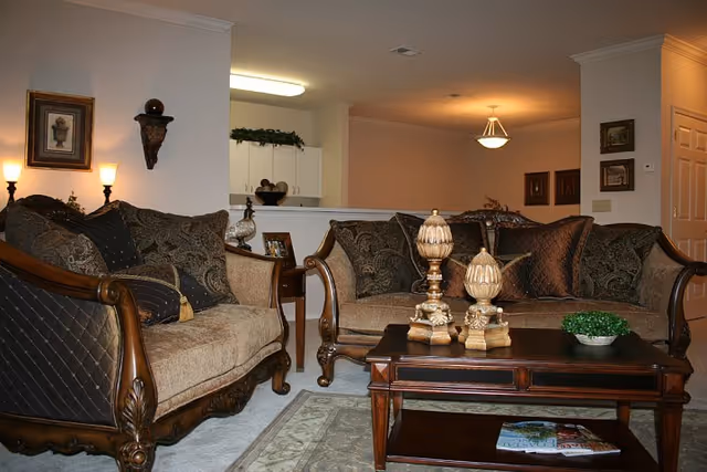Elegant living room with two carved wooden sofas, patterned cushions, a wooden coffee table topped with decorative urns, and a view into the adjacent kitchen/dining area.