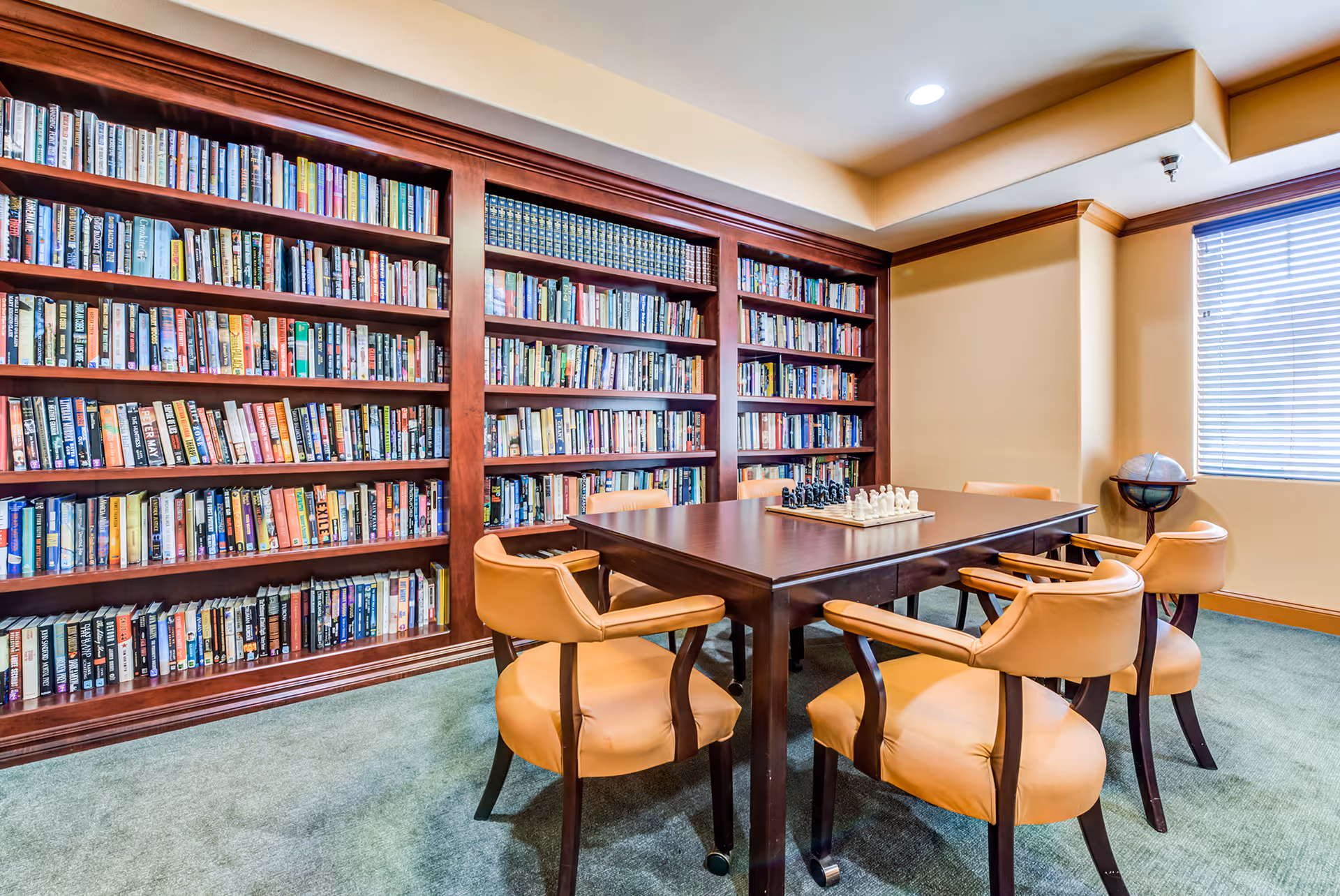 A cozy library room with a large wooden bookshelf filled with books along one wall. In the center, there is a dark wooden table with a chess set on it, surrounded by six tan leather chairs. A window with blinds allows natural light into the room, and a globe stands in the corner.