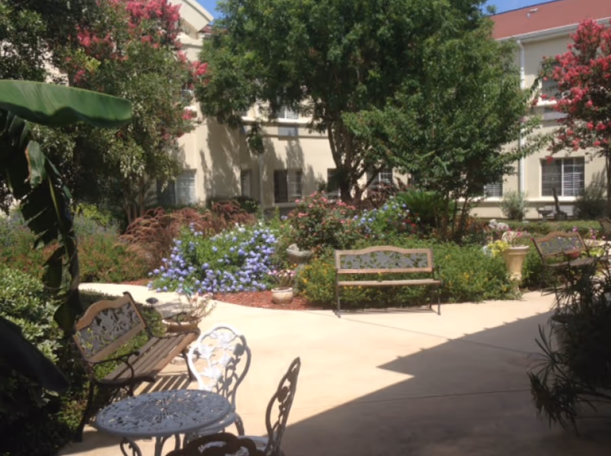 A sunny outdoor garden area at Esplanade Gardens featuring several benches and a small round metal table with chairs. The garden is lush with various green plants, trees, and colorful flowers including purple and pink blooms. The building with windows and a red roof is visible in the background.