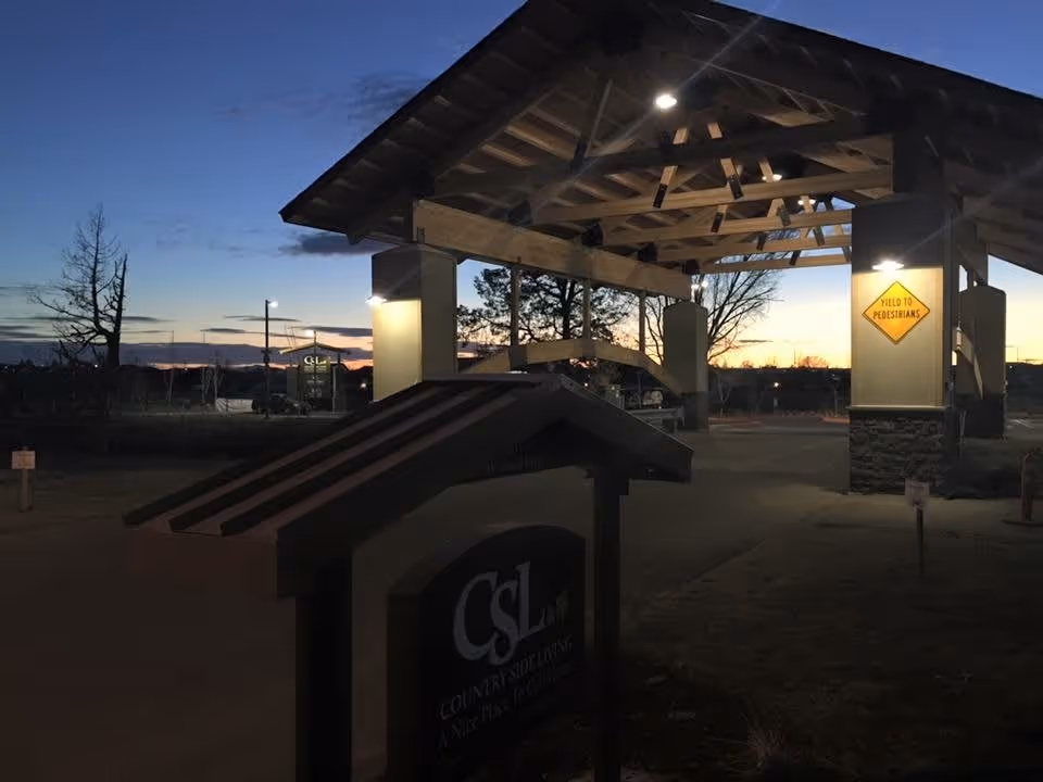 Entrance area of Country Side Living - Memory Care facility at dusk, featuring a covered drop-off area with wooden beams and stone pillars, illuminated by lights. A sign in the foreground displays the facility's name and logo. A yellow traffic sign on one pillar reads 'Yield to Pedestrians'. Trees and a darkening sky are visible in the background.