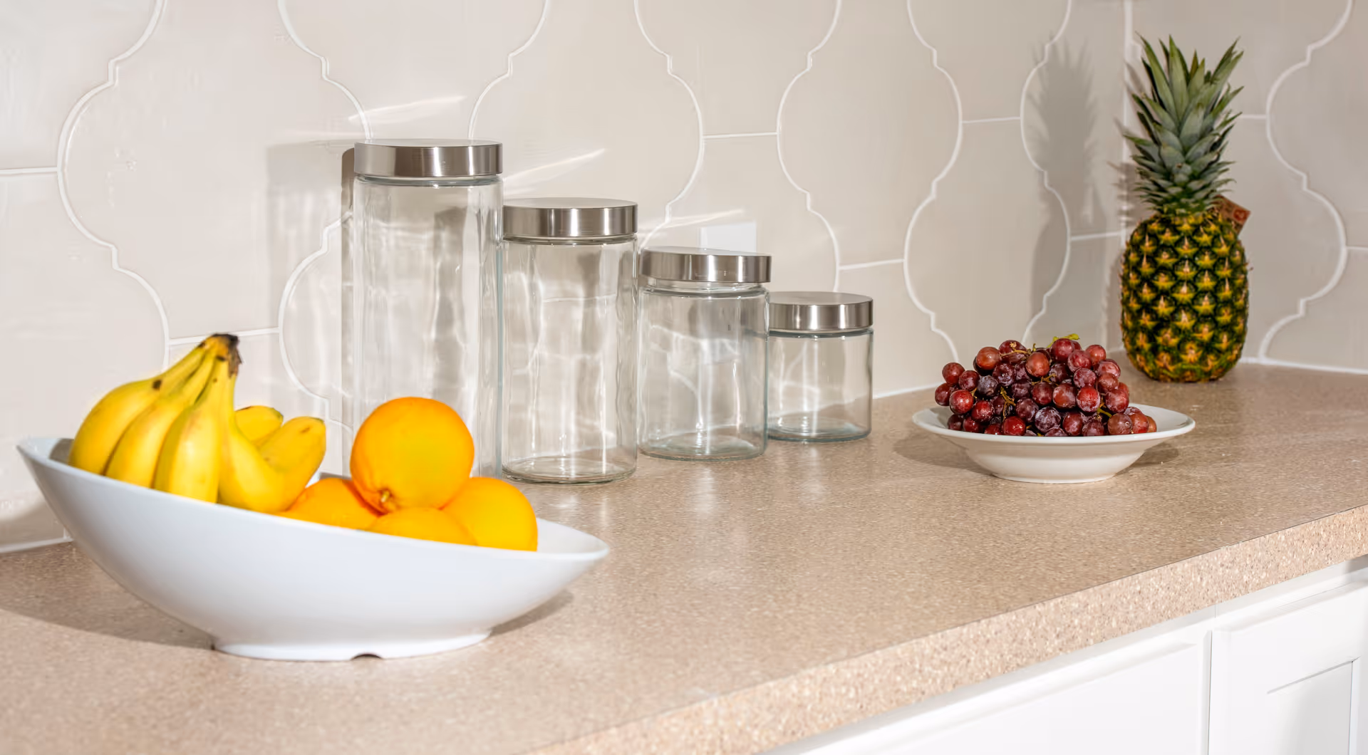 A kitchen countertop with a white bowl containing bananas and oranges, four empty glass jars with metal lids arranged in descending size, a white plate with red grapes, and a whole pineapple in the background against a white tiled backsplash.