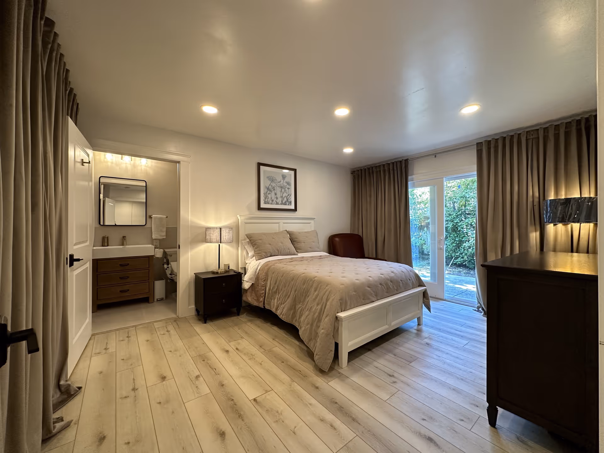 A cozy bedroom with a white wooden bed frame and beige bedding. There is a nightstand with a lamp next to the bed, a dark wooden dresser with a lamp on the right side, and a brown armchair in the corner. The room has light wood flooring and beige curtains covering large glass doors that open to a garden outside. An open door reveals a bathroom with a vanity, sink, mirror, and towel rack.
