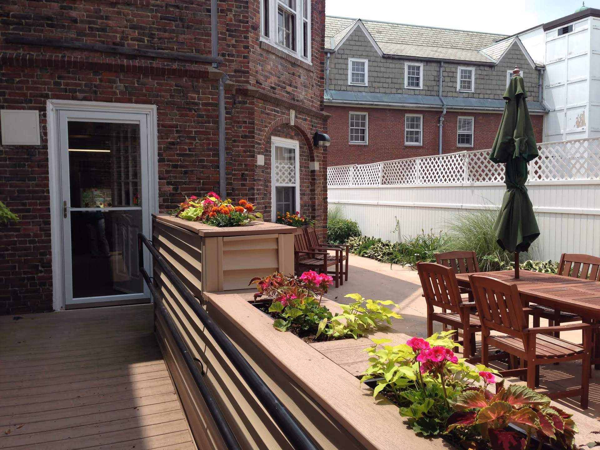 Outdoor courtyard patio with wooden tables and chairs, umbrellas, flower-filled planters, and a brick building with a white fence.