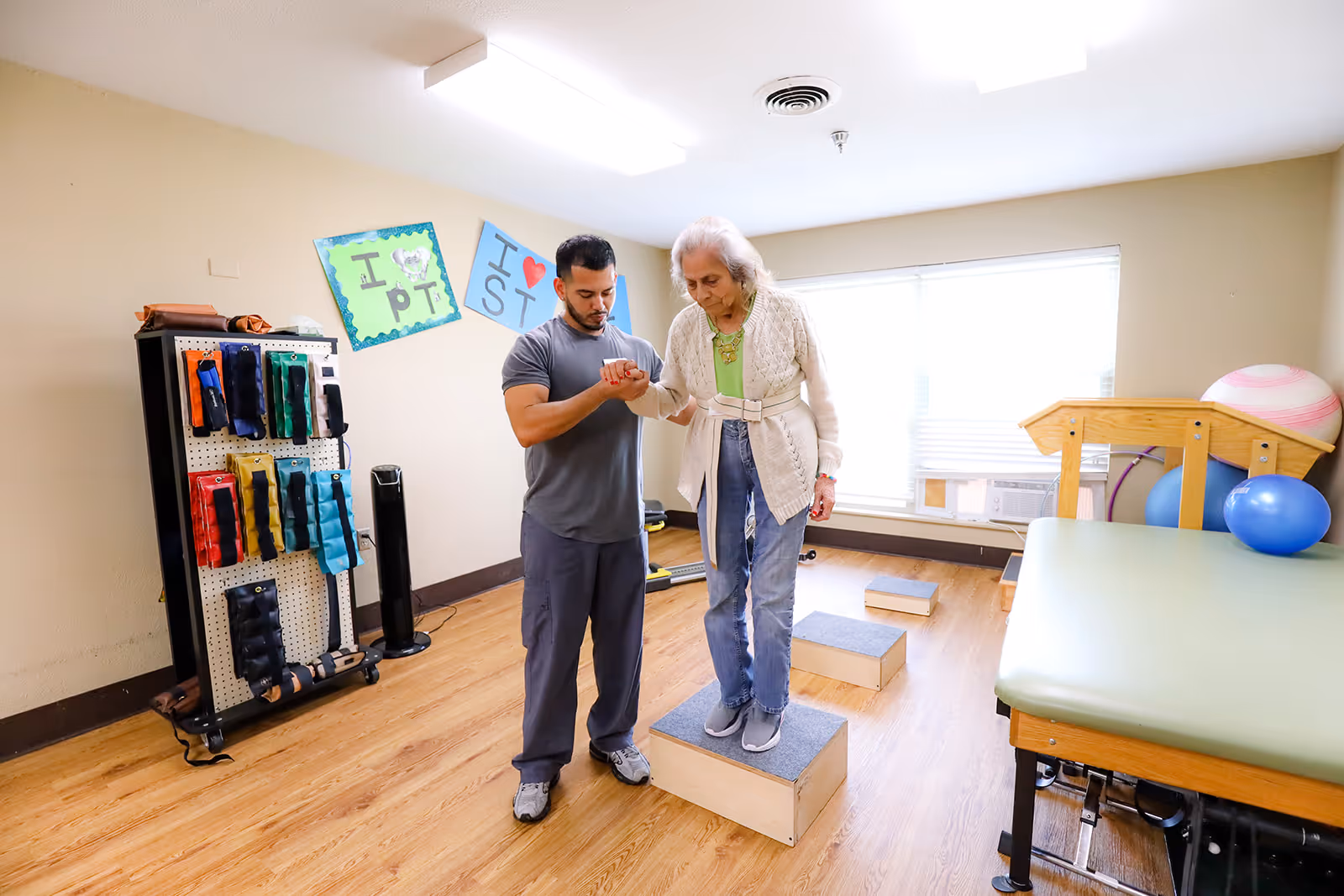 A male physical therapist assists an elderly woman as she steps onto a wooden block in a rehabilitation room. The room has wooden flooring, exercise equipment including resistance bands on a rack, therapy balls, and a padded therapy table. There are two colorful posters on the wall with messages related to physical therapy.