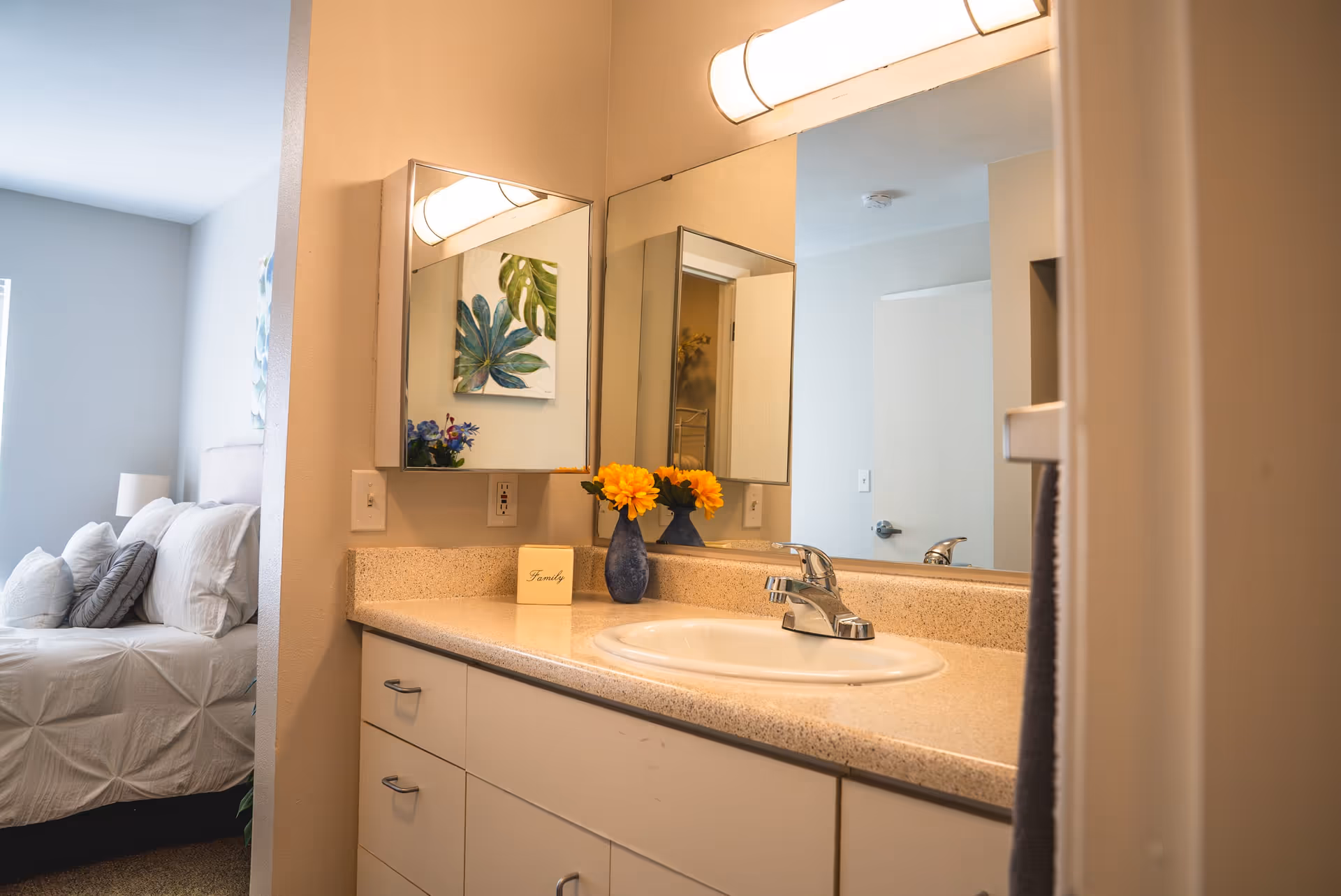 A bathroom vanity with a sink, countertop, and two mirrors above it. A blue vase with yellow flowers and a small block with the word 'Family' are placed on the countertop. In the background, a bedroom with a bed, white bedding, and pillows is partially visible.