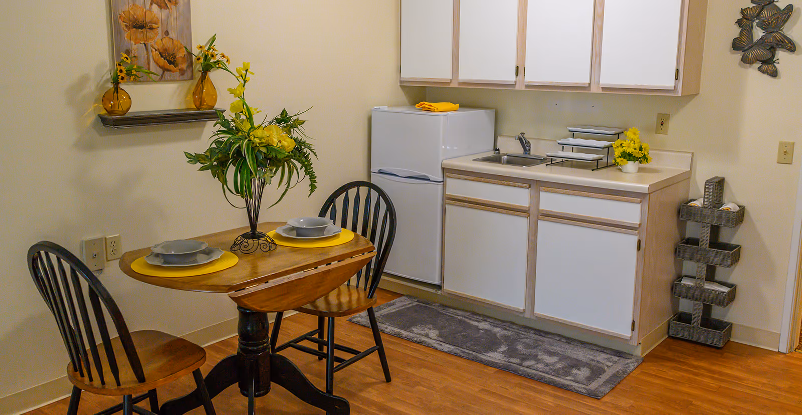A small kitchen area with white cabinets, a white refrigerator, and a sink. In front of the kitchen is a wooden table set for two with yellow placemats, gray plates, and a floral centerpiece. The walls are decorated with a painting of flowers and a metal butterfly wall art. There is a gray rug on the wooden floor and a small wicker shelf holding rolled towels.