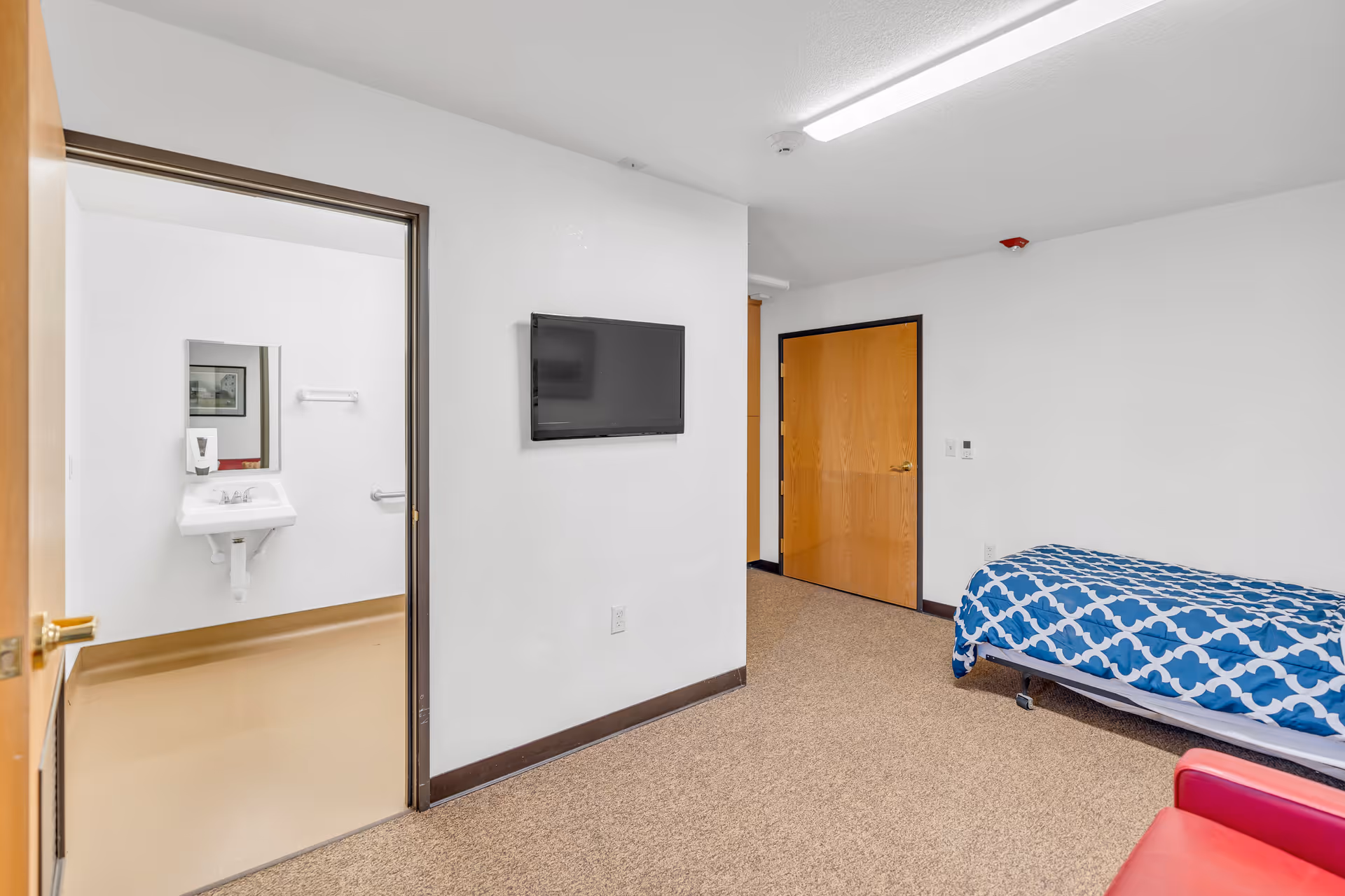 A simple room with a single bed covered in a blue and white patterned blanket, a red chair in the foreground, a wall-mounted flat screen TV, and an open door leading to a bathroom with a sink, mirror, and soap dispenser.