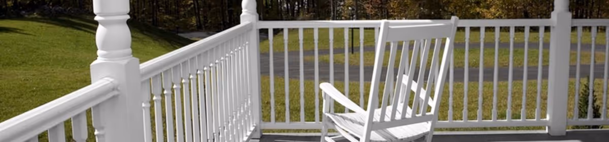 A white wooden rocking chair sits on a porch with white railings overlooking a grassy lawn, road, and trees.