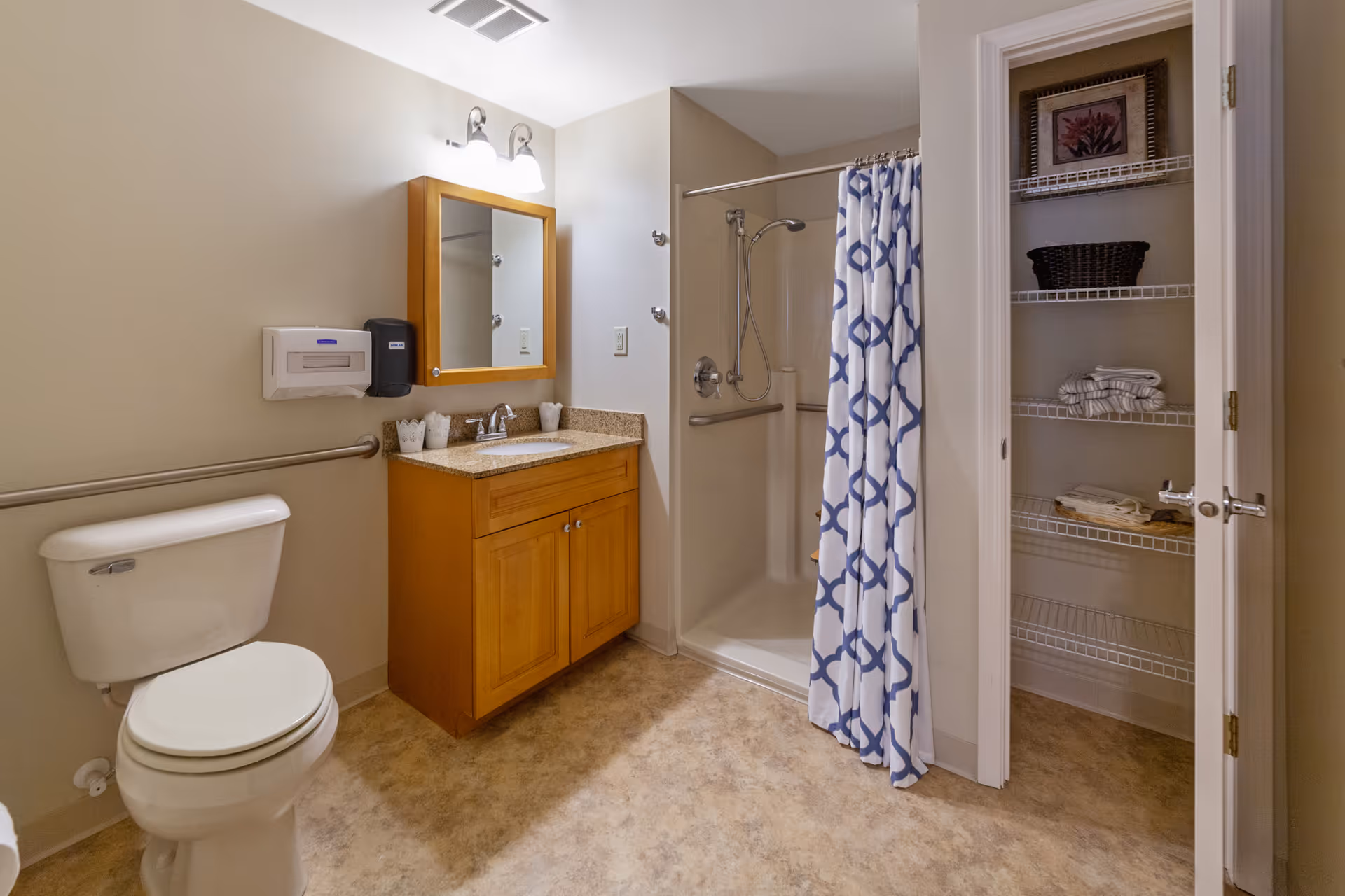A clean and well-lit bathroom featuring a white toilet, a wooden vanity with a granite countertop and a sink, a mirror with two lights above it, a shower with a blue and white patterned curtain, and an open closet with shelves holding towels, a basket, and a framed picture.