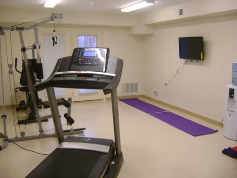 A small fitness room with a treadmill, multi-exercise gym machine, purple exercise mat on the floor, wall-mounted TV, and a water cooler in the corner.