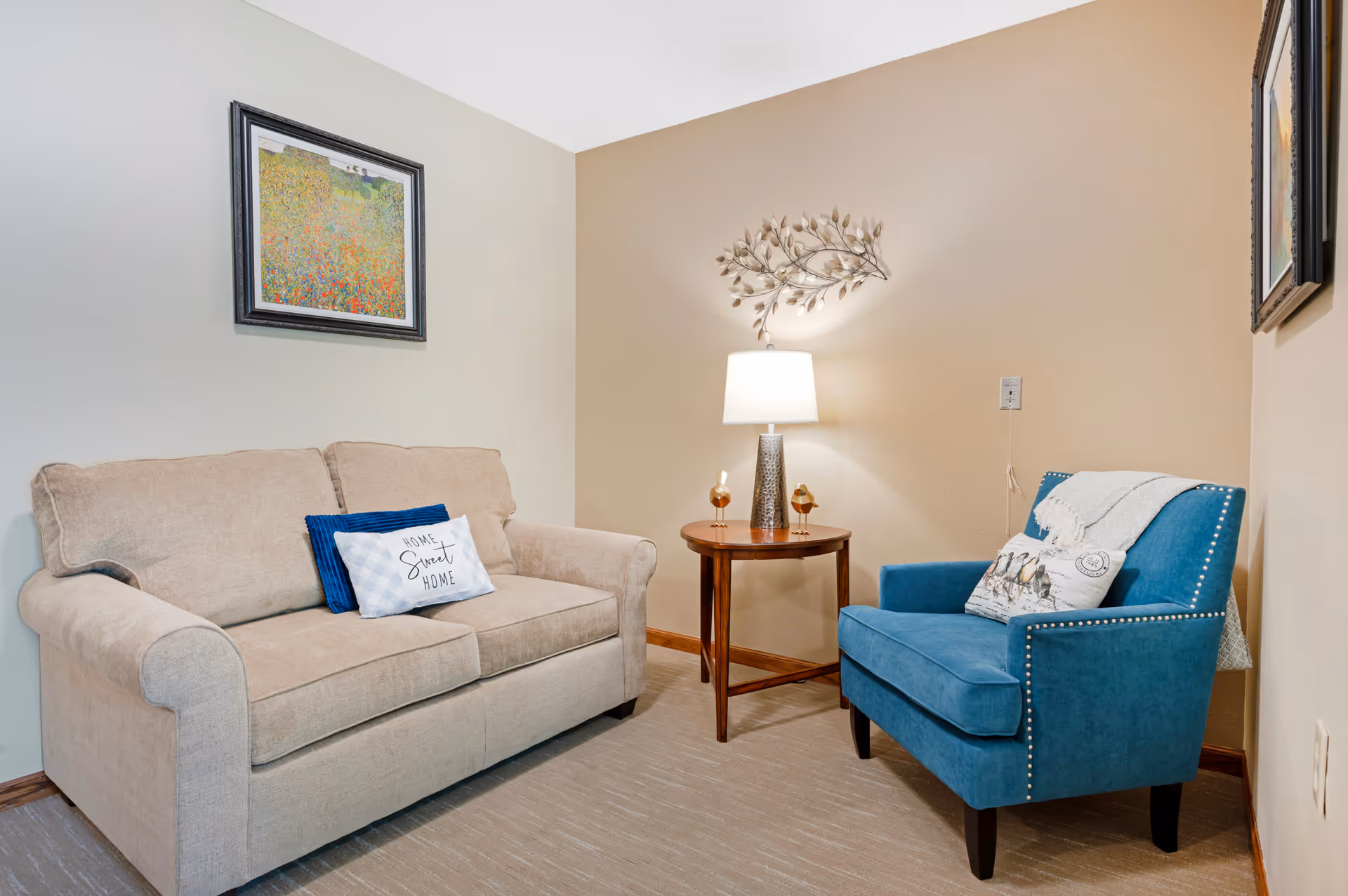 A cozy seating area with a beige loveseat adorned with two decorative pillows, one reading 'Home Sweet Home,' and a blue armchair with a throw blanket and pillow. Between the seats is a wooden side table with a lamp and two small decorative items. The walls are painted in two tones, beige and off-white, with framed artwork and a metal leaf wall decoration.
