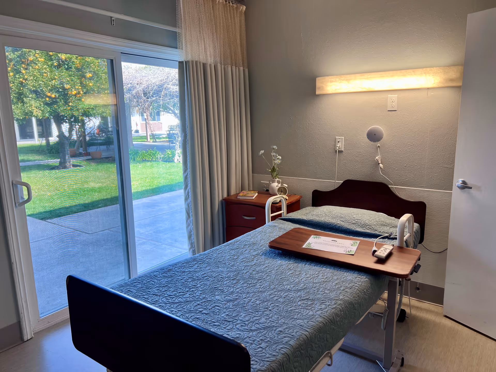 A small bedroom in a senior living facility with a single bed covered with a blue quilt. The bed has white safety rails and a wooden overbed table with a remote control and a paper on it. Next to the bed is a wooden nightstand with a small vase of flowers and a book. A large sliding glass door with curtains opens to a green outdoor area with trees and a walkway. The room has a wall-mounted light above the bed and a partially open door on the right.