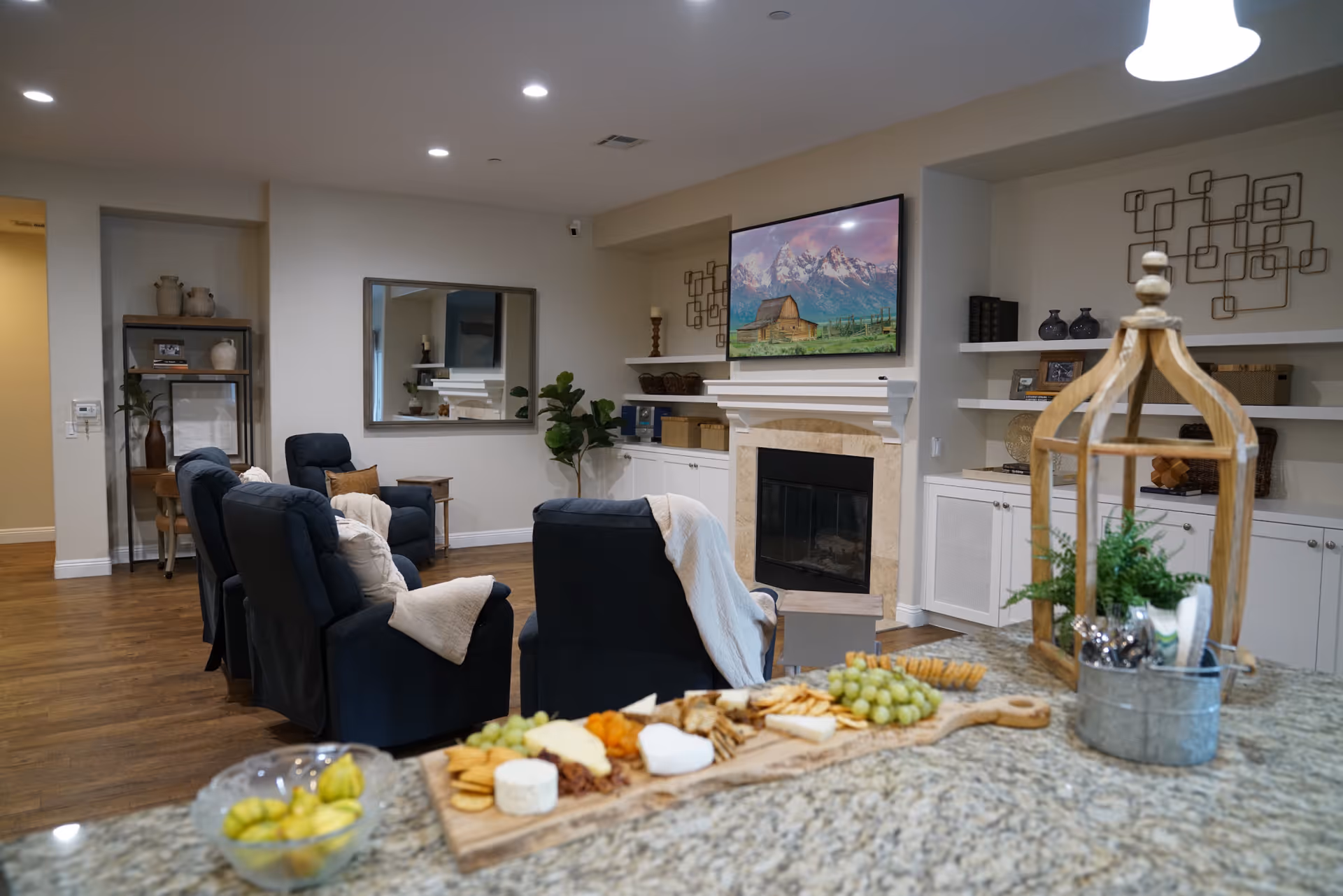 Open communal living room with recliner chairs facing a fireplace and wall-mounted TV, and a granite countertop with a cheese board in the foreground.