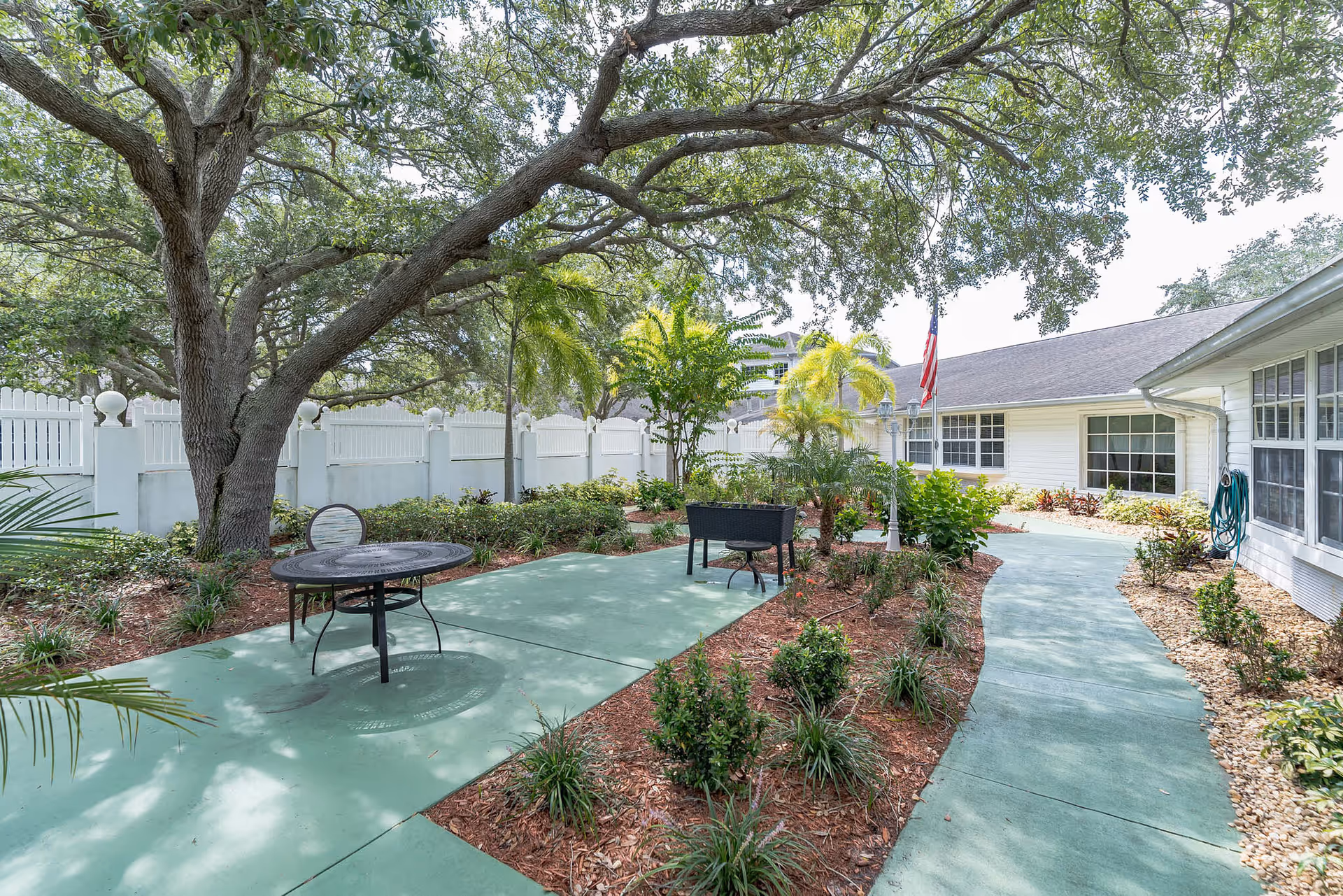 Outdoor garden area at The Landmark with a large tree providing shade, a round metal table with chairs, a pathway surrounded by plants and shrubs, and a white building with multiple windows in the background. An American flag is visible near the building.