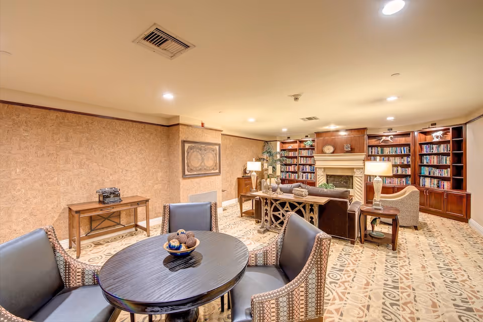 Cozy communal lounge and library with bookshelves, a fireplace, a round table and upholstered chairs.