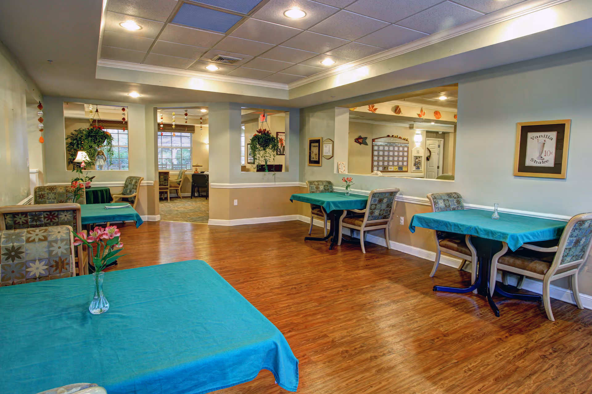 Interior view of a senior living facility dining area with several tables covered in teal tablecloths, each accompanied by cushioned chairs. The room has wooden flooring, light-colored walls, ceiling lights, and decorative plants hanging near the windows. A framed picture on the wall reads 'Vanilla Shake 10¢'.