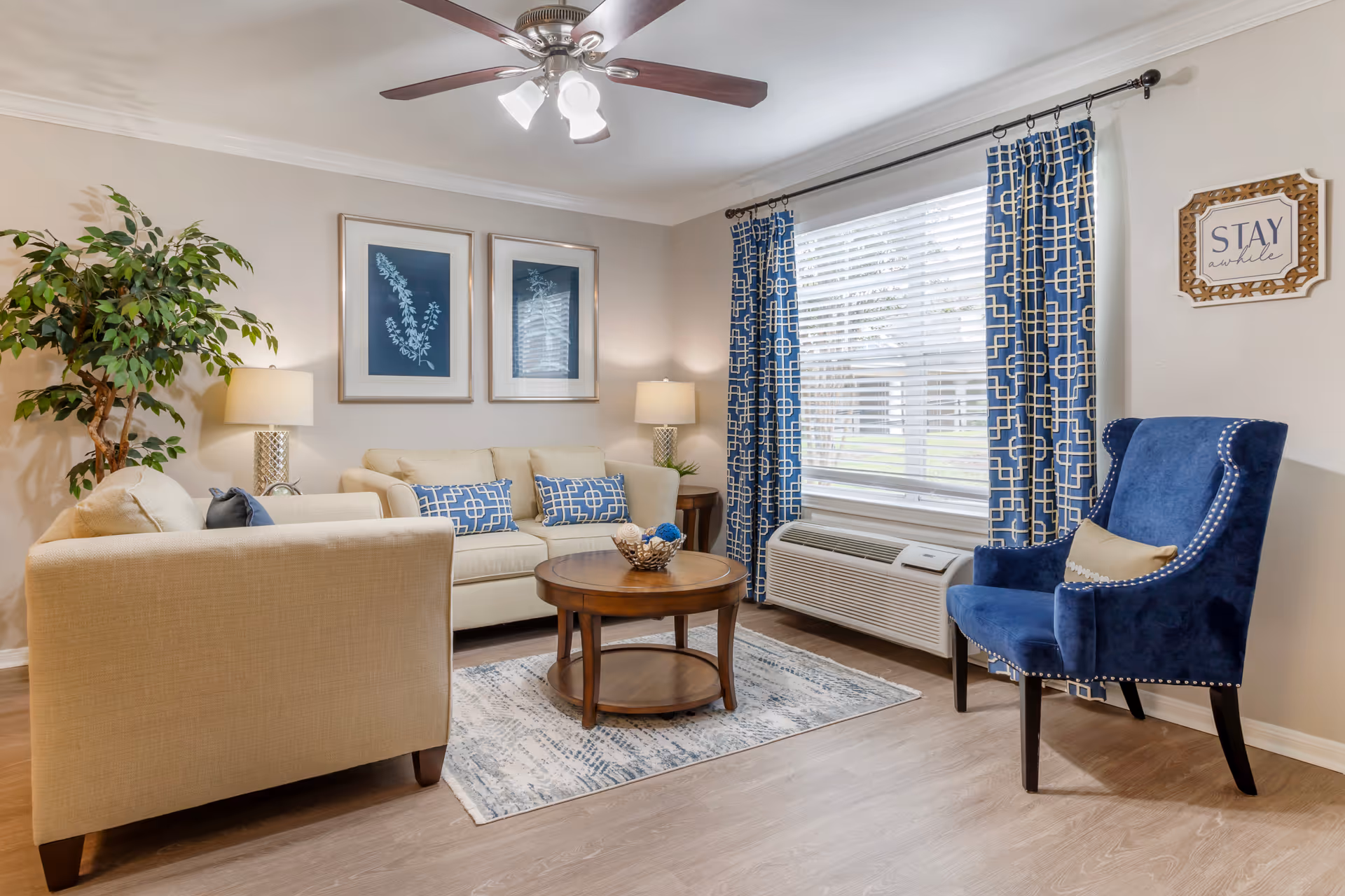 A cozy living room with beige sofas adorned with blue patterned pillows, a round wooden coffee table on a light blue and white rug, a blue upholstered armchair with a beige pillow, two framed botanical prints on the wall, blue patterned curtains on a window with white blinds, two table lamps, a ceiling fan with lights, and a potted plant in the corner. A wall sign reads 'STAY awhile'.