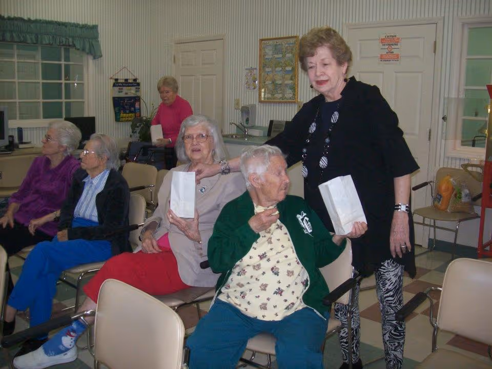A group of elderly women seated in a room with chairs arranged in rows. One woman stands beside another who is eating a snack from a white paper bag. The room has a checkered floor, a window with a green valance, and various items on the walls and counters.