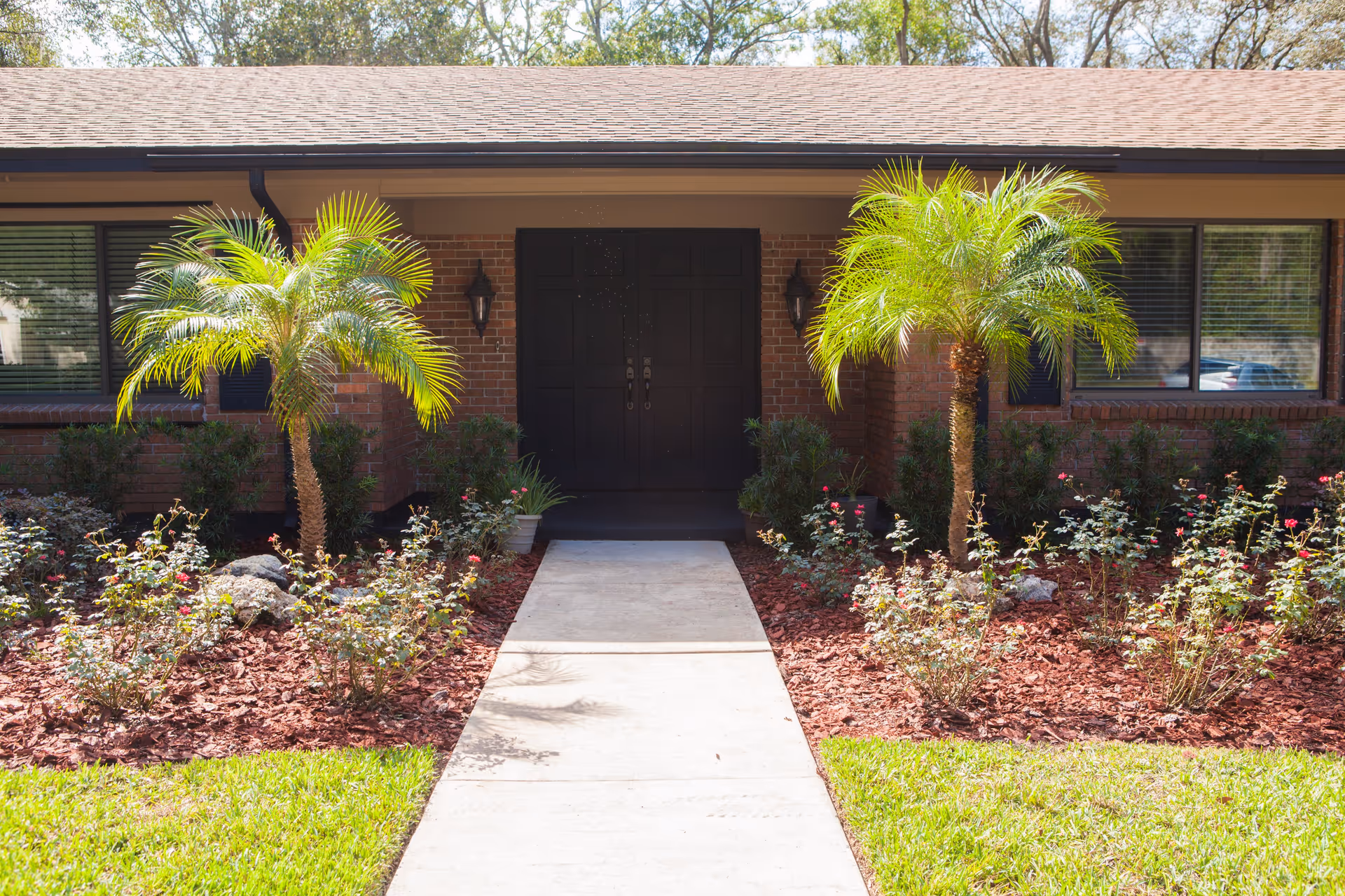 Front entrance of a brick building with a dark double door, flanked by two wall-mounted lantern lights. A concrete pathway leads to the door, bordered by landscaped garden beds with small palm trees, rose bushes, and red mulch. Green grass surrounds the pathway and garden beds.