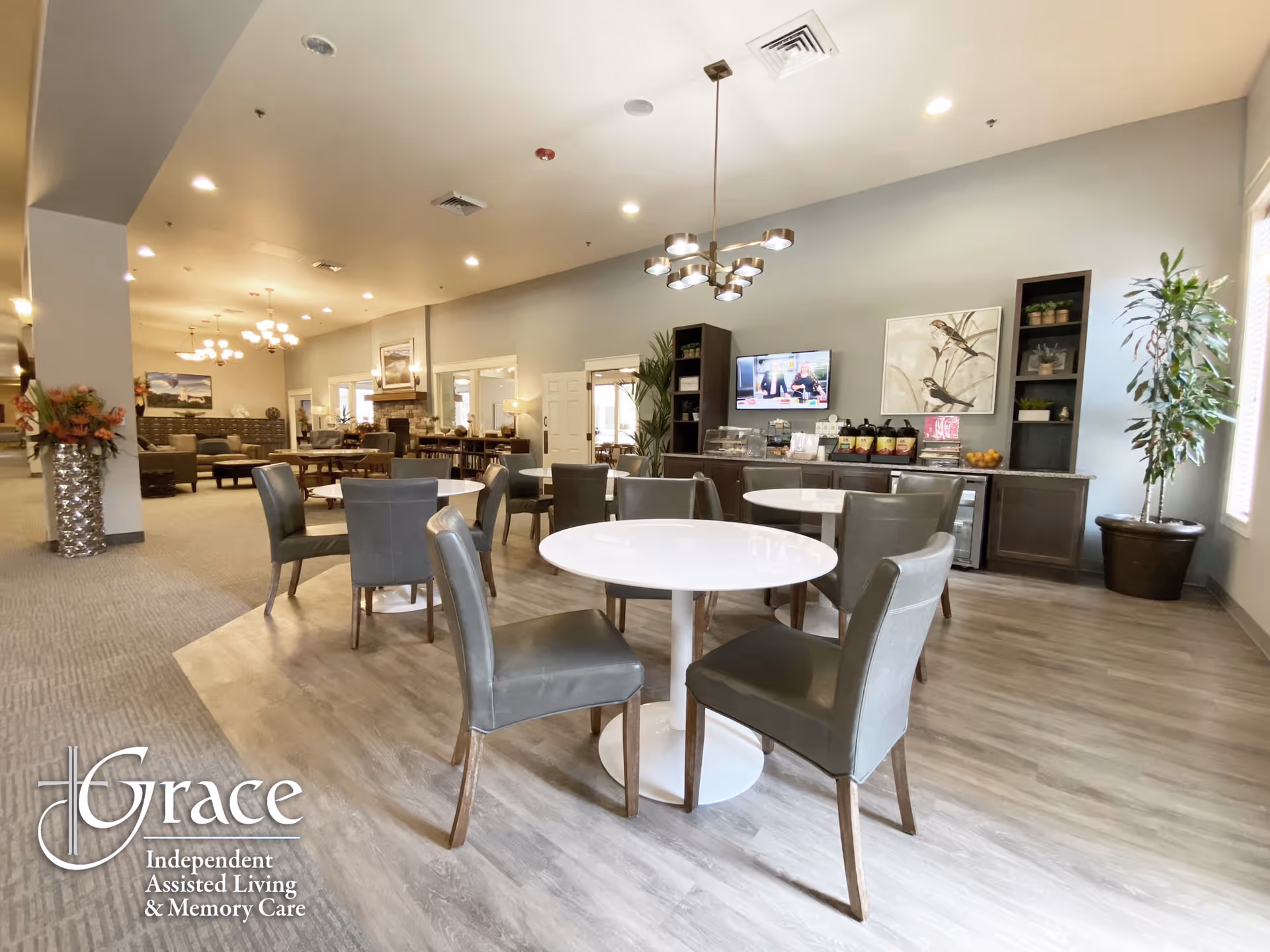 A spacious and well-lit dining area in an assisted living facility with several round white tables surrounded by gray chairs. The room features modern light fixtures, a television mounted on the wall, decorative plants, and a sideboard with refreshments. The flooring is a combination of wood and carpet, and the overall decor is contemporary and welcoming.