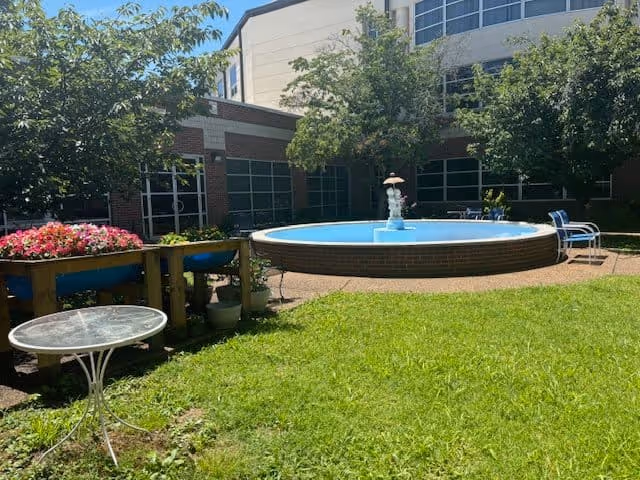 Outdoor courtyard area with a circular water fountain in the center, surrounded by green grass, trees, and some chairs. There is a small round glass table on the left side and a flower planter with pink flowers. The building with large windows is visible in the background.