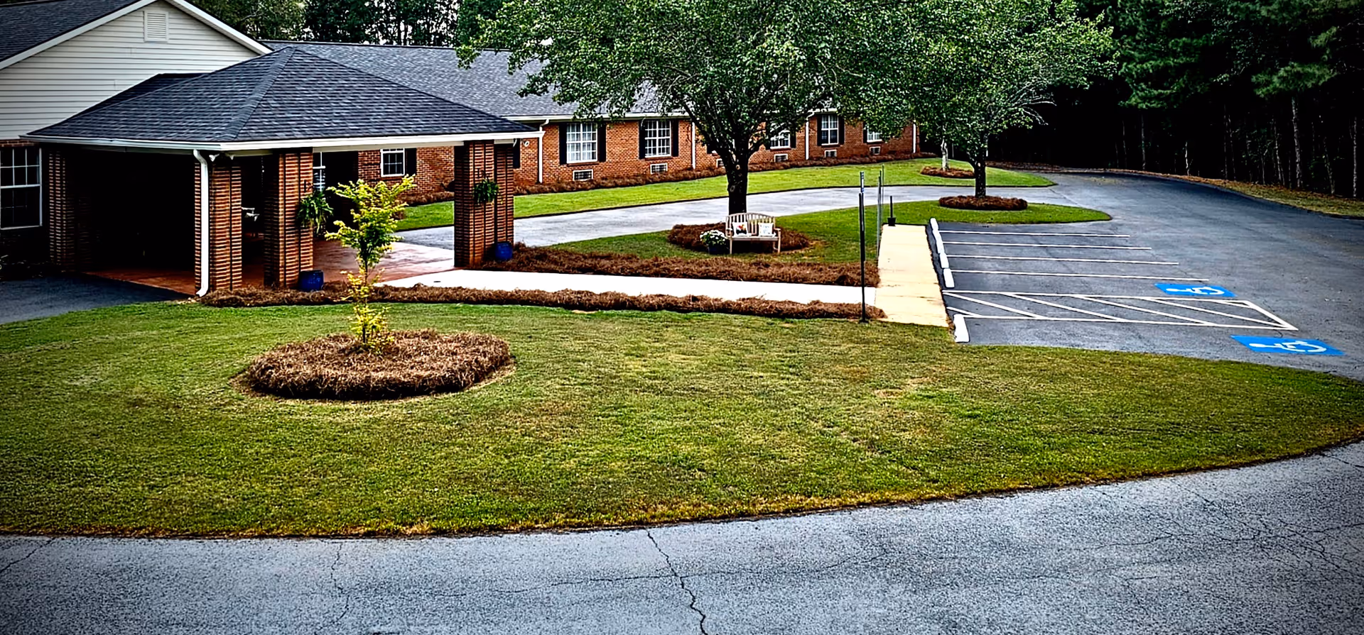Front exterior of a single-story brick memory care facility with a covered entrance, circular driveway, lawn with trees and benches, and a parking area with accessible spaces.