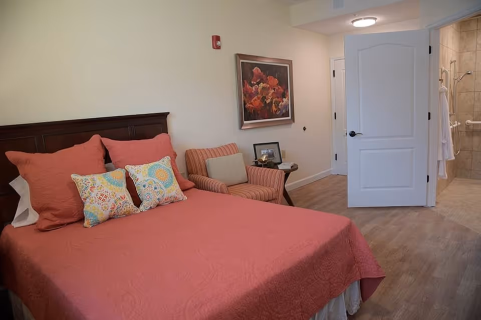A bedroom with a bed covered in a coral-colored quilt and matching pillows, two decorative patterned pillows, a striped armchair with a beige cushion, a small side table with a framed photo and decorative items, a floral painting on the wall, and an open door leading to a bathroom with a tiled shower and a white robe hanging inside.