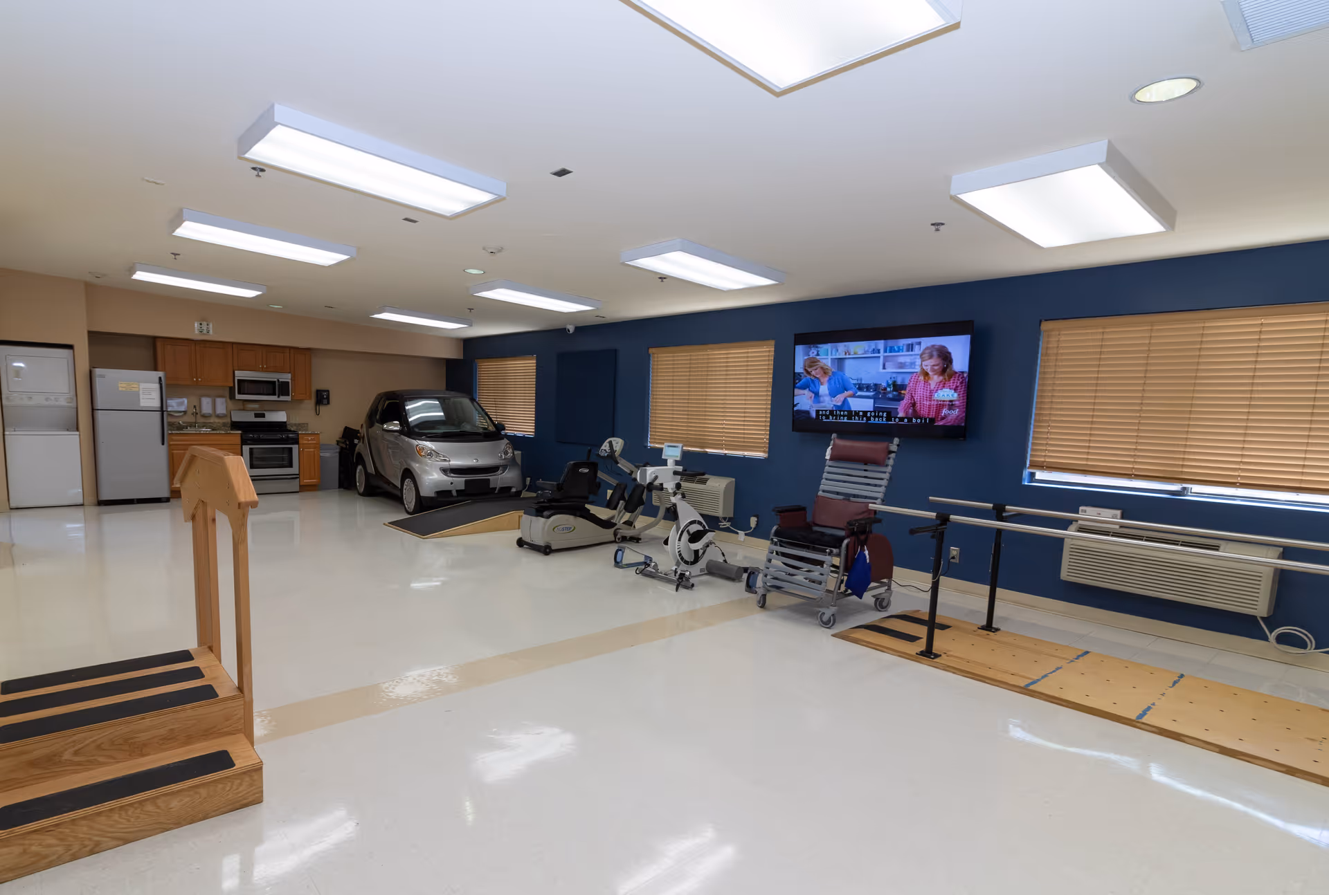 A spacious rehabilitation room with a small car on a ramp, exercise equipment, a wheelchair, parallel bars for walking practice, a wooden staircase for physical therapy, a kitchenette with a refrigerator, microwave, and stove, and a wall-mounted TV showing two people in a kitchen setting. The room has bright overhead lighting and windows with closed blinds.