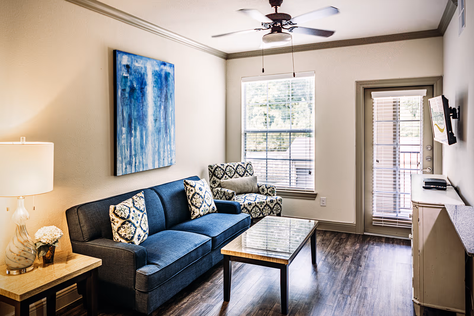Bright living room with a blue sofa, patterned armchair, coffee table, and a window and door leading to a balcony.