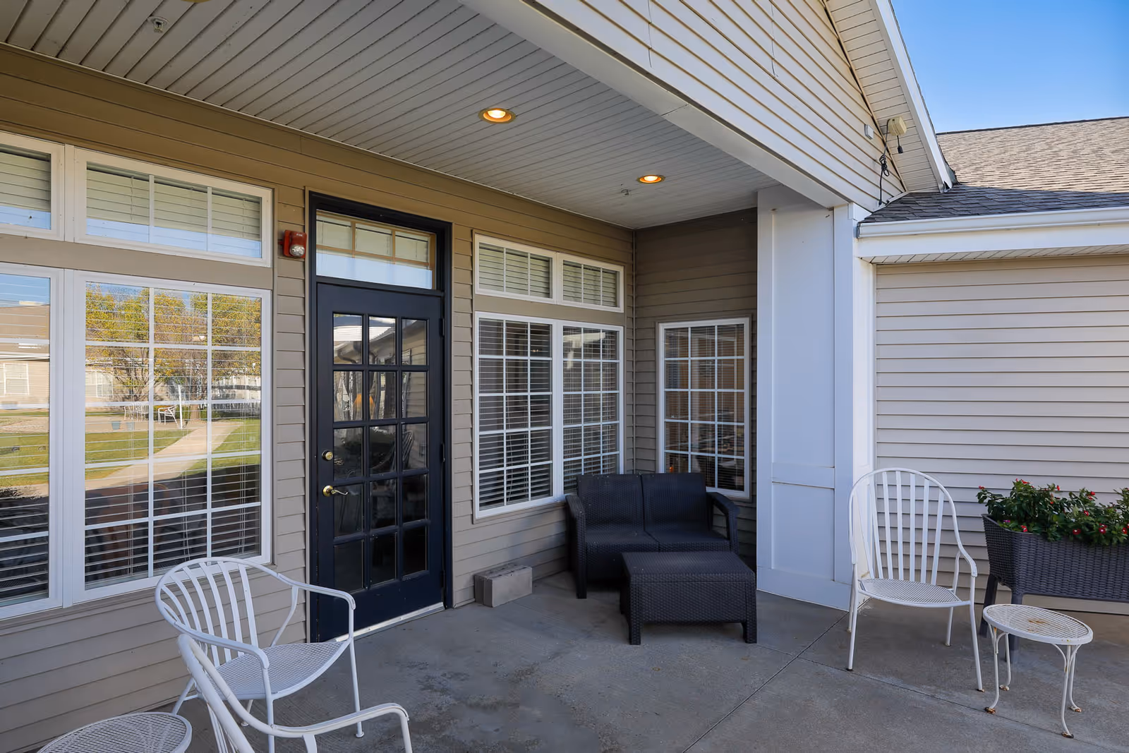 Covered outdoor patio area with beige siding walls, a black door with glass panels, multiple windows with white blinds, two white metal chairs, a small white metal table, a black wicker loveseat with a matching ottoman, and a planter with flowers.