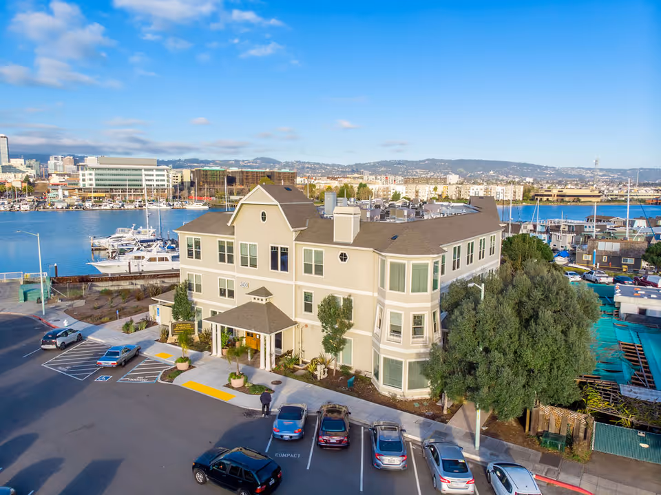 A three-story beige building with multiple windows and a small covered entrance, located next to a marina with boats docked in the water. Several cars are parked in front of the building, and there are trees and landscaping around the property. The background shows a cityscape with hills under a blue sky with some clouds.