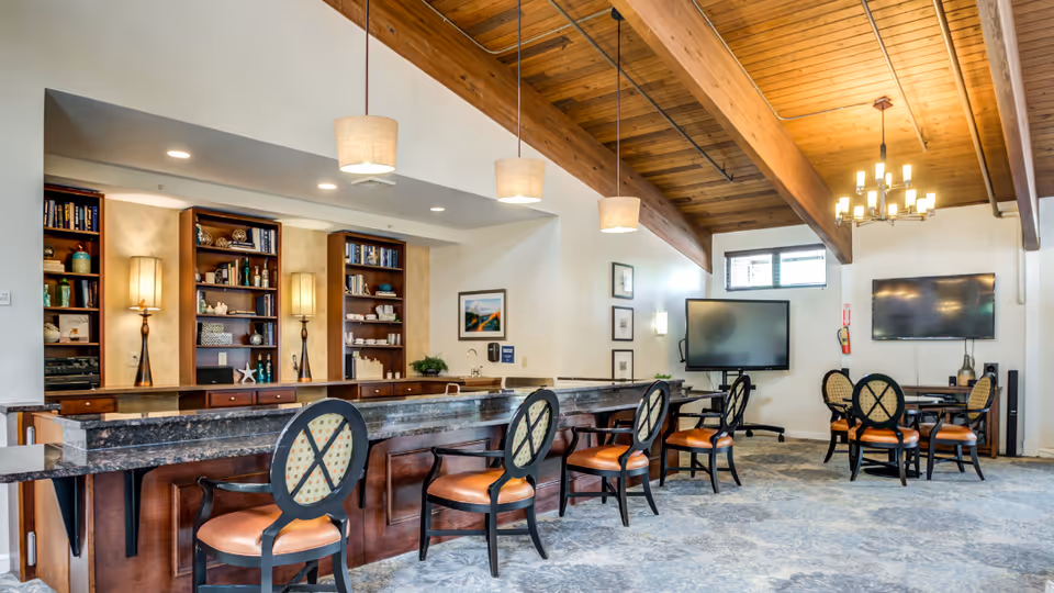 A spacious common area with a high wooden ceiling featuring exposed beams. There is a long granite countertop with six cushioned chairs lined up in front of it. Behind the counter are built-in wooden shelves with decorative items and two tall lamps. The room also has a carpeted floor, two large flat-screen TVs mounted on the walls, a round table with four chairs, and modern pendant and chandelier lighting.