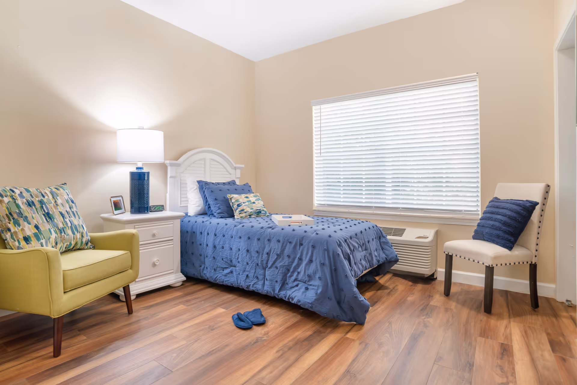 A cozy bedroom with a single bed covered in a blue quilt and matching pillows. Next to the bed is a white nightstand with a blue lamp, a framed photo, and an alarm clock. A green armchair with patterned cushions is on the left, and a beige chair with a blue pillow is on the right near a large window with white blinds. The floor is wooden, and a pair of blue slippers is placed near the bed.
