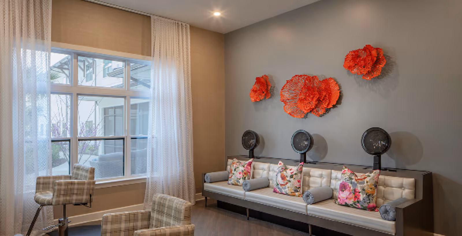 A modern salon area with three vintage hair dryers mounted on a long cushioned bench with floral pillows. There are two plaid salon chairs in front of the bench and a large window with sheer white curtains letting in natural light. The wall behind the bench is decorated with three large red floral wall sculptures.