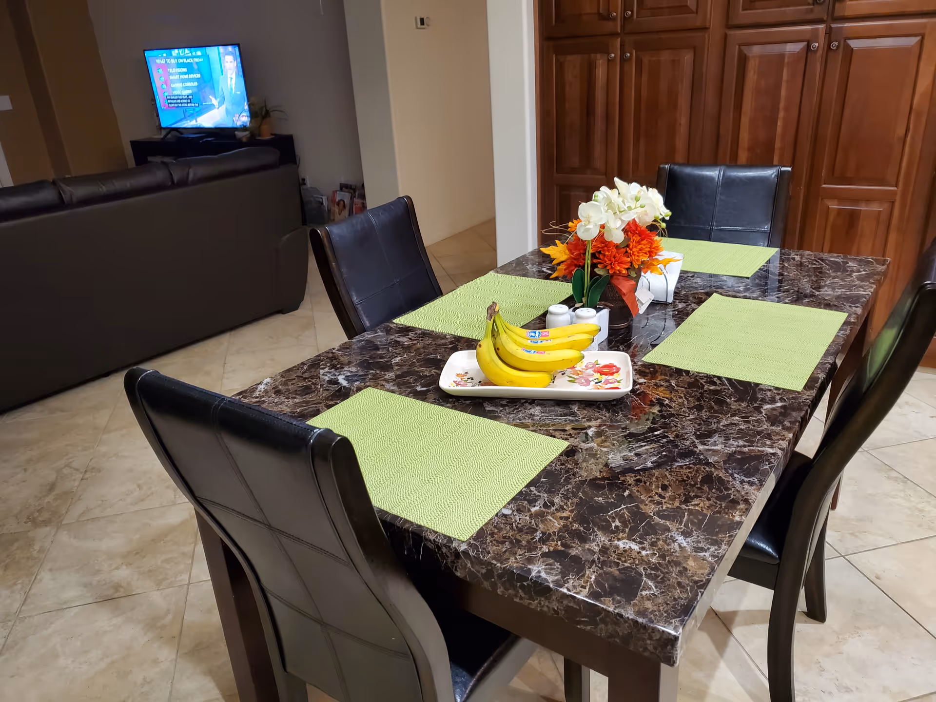 A dining area with a dark marble table set with four green placemats and a centerpiece of flowers and bananas on a tray. Four black leather chairs surround the table. In the background, there is a living room area with a dark sofa and a television mounted on a stand.