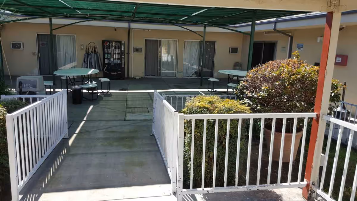 Outdoor patio area at Medical Center Convalescent Hospital with concrete walkway, white metal railings, round tables with attached benches, vending machines, and potted plants under a green canopy.