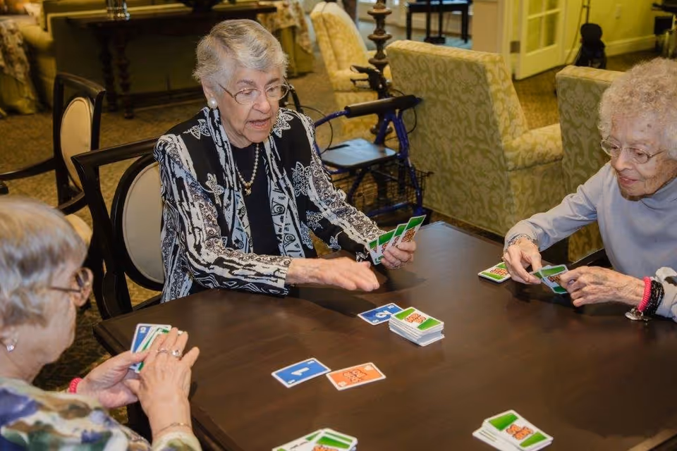 Three elderly women sitting around a table playing a card game in a cozy room with patterned armchairs and a walker in the background.