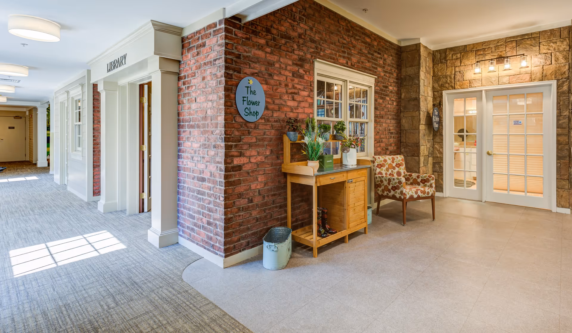Interior hallway of Arden Courts - ProMedica Memory Care Community (Avon) featuring a cozy corner with a wooden table decorated with plants, a floral patterned armchair, and a sign reading 'The Flower Shop' on a brick wall. To the left, there is a door labeled 'Library' and a carpeted corridor with natural light coming through windows.