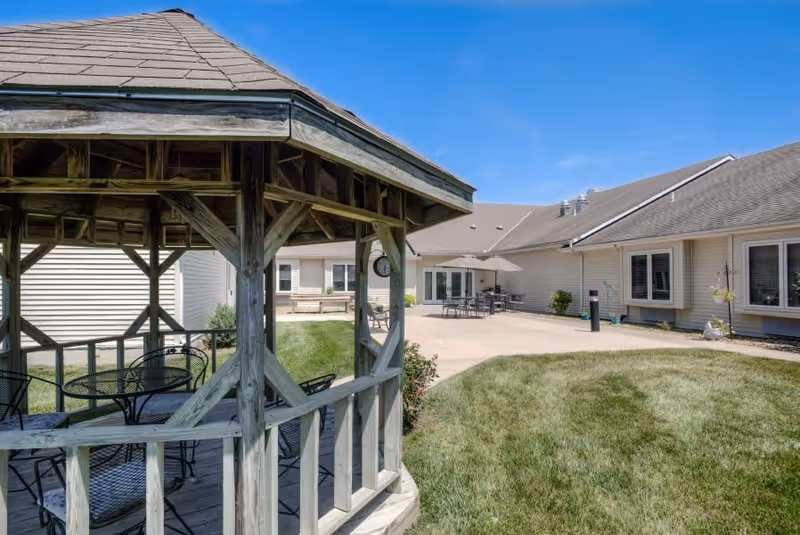 Outdoor patio area at Vintage Park at Baldwin City featuring a wooden gazebo with metal chairs and a table inside, a grassy lawn, and a paved area with tables, chairs, and umbrellas next to a beige building under a clear blue sky.