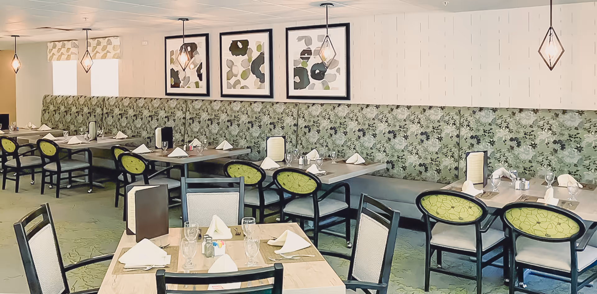 A dining room with multiple set tables, green patterned banquette seating, black chairs with green cushions, and framed artwork on the wall.