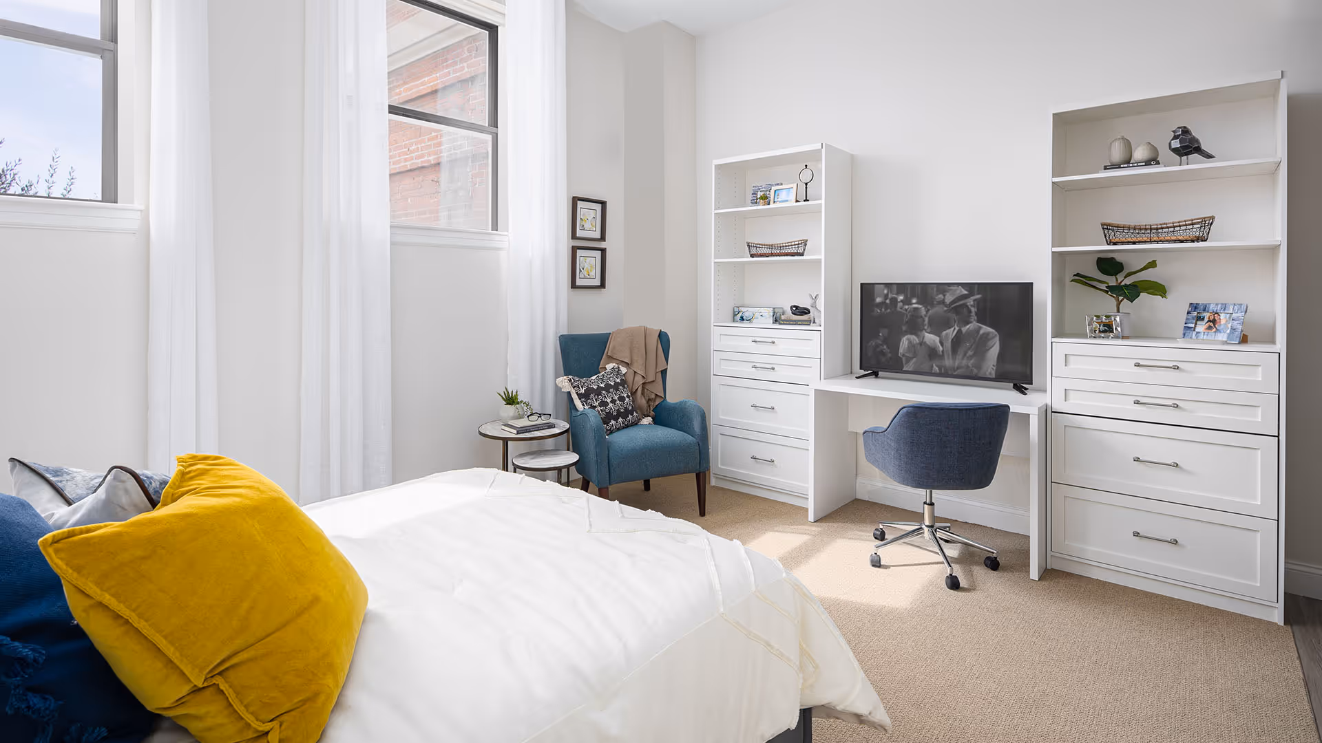 A bright bedroom with a bed featuring yellow and blue pillows, a blue armchair with a throw blanket and pillow, a small round side table with a plant and glasses, two large windows with white curtains, and a white built-in desk with a TV and a blue swivel chair. There are two white shelving units with decorative items and framed photos.