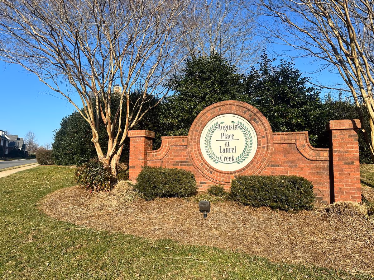 A brick entrance sign that reads "Augusta Place at Laurel Creek" set in landscaped grounds with shrubs, trees, and a clear blue sky.