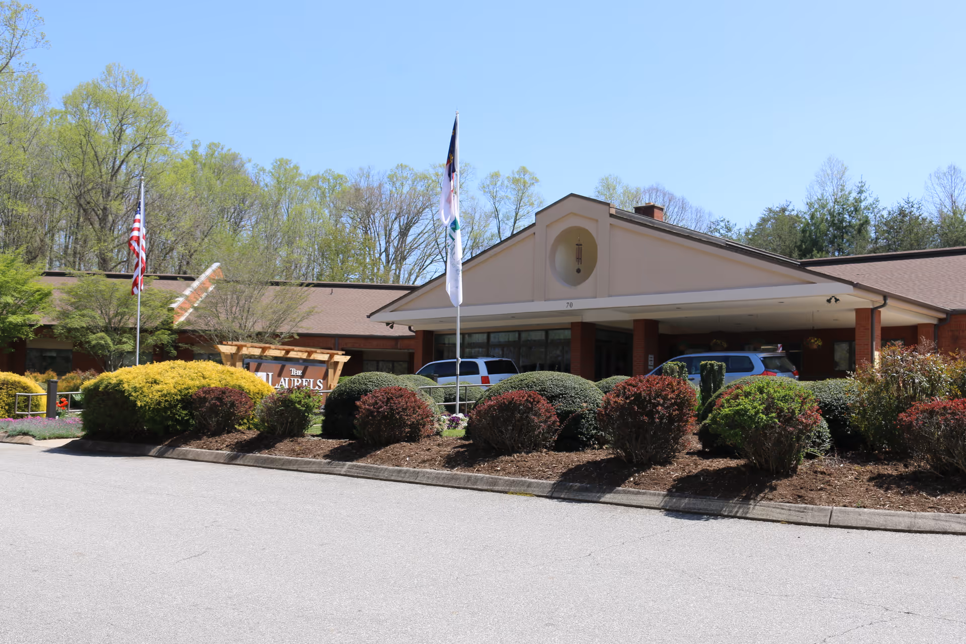 Exterior view of The Laurels of GreenTree Ridge facility showing the main entrance with a covered drop-off area, surrounded by well-maintained bushes and landscaping. Two flagpoles with flags are visible in front of the building, and several vehicles are parked near the entrance. Trees and a clear blue sky are in the background.