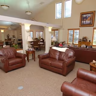 A spacious living room area in a senior living facility with brown leather armchairs and sofas arranged around small wooden tables. The room has beige carpeting, large windows letting in natural light, and high ceilings with ceiling lights. There are decorative items on tables and a framed picture on the wall.