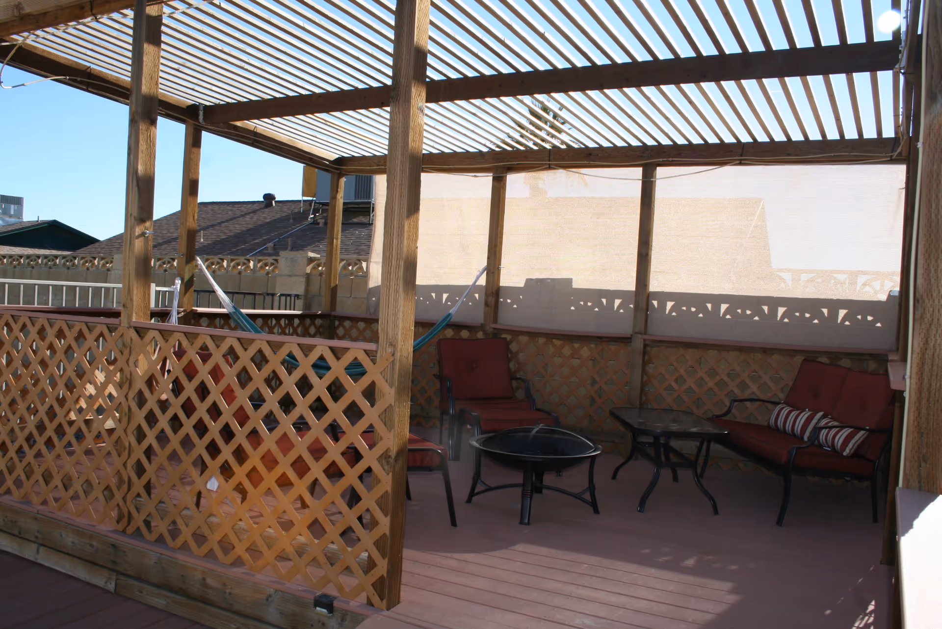 Wooden pergola-covered outdoor deck with lattice railing, red cushioned seating, glass tables and a hammock.