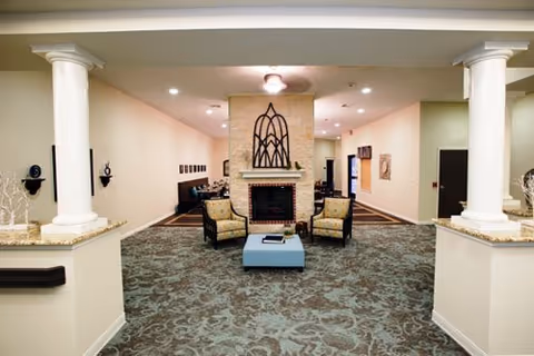 Interior view of a common area in a senior living facility featuring a central fireplace with decorative metal art above it, two armchairs, and a blue ottoman. The space is carpeted with a patterned design and flanked by two white columns on either side. The walls are light-colored with some framed artwork and lighting fixtures on the ceiling.